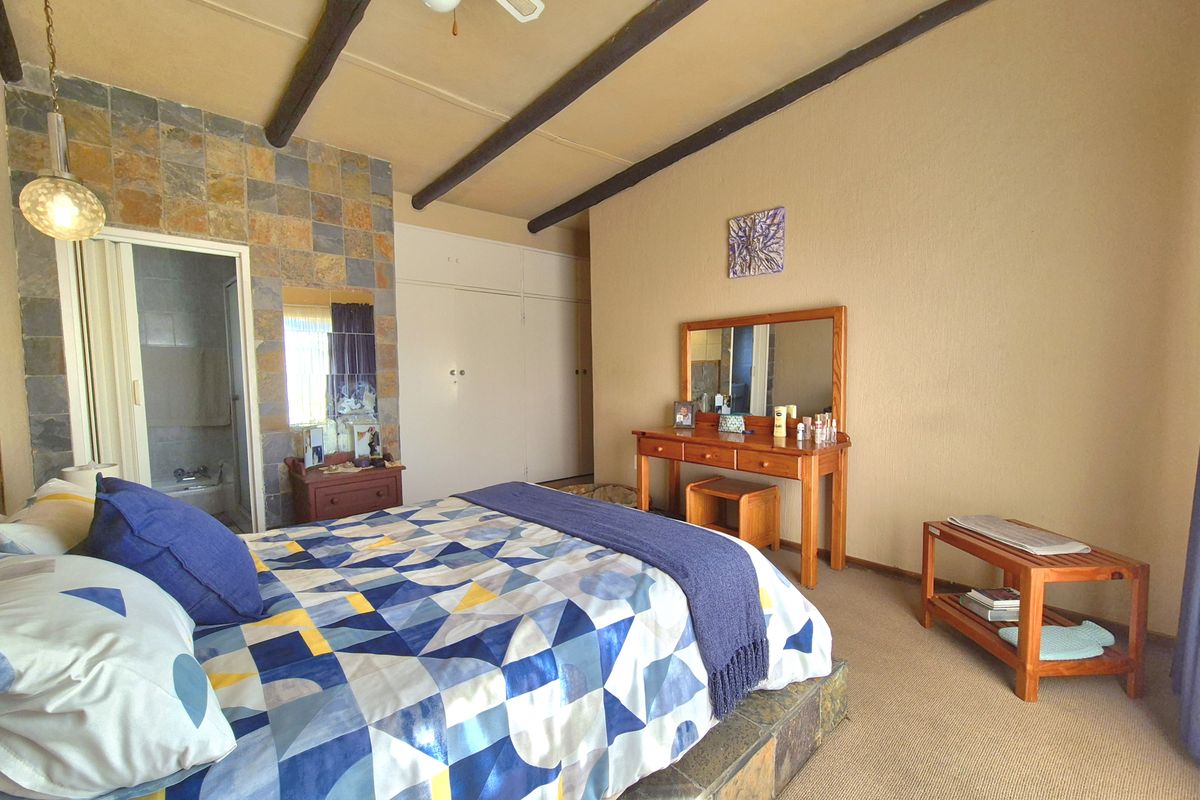 Spacious bedroom featuring exposed timber ceiling beams, a stone-tiled feature wall, built-in cupboards, and a raised stone-tiled bed platform with en-suite access.