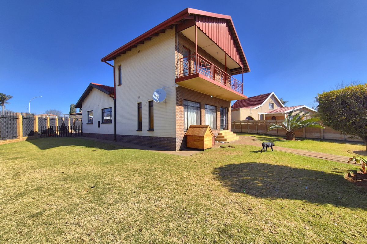 Rear view of a two-story brick house featuring a balcony, metal roofing, and a spacious garden with a paved walkway.