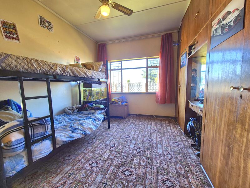 A bedroom featuring built-in wooden cupboards, a ceiling fan, and a large window with security bars, finished with patterned carpeting.