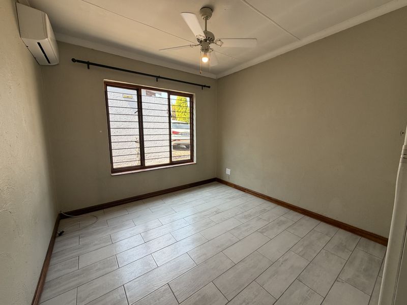 A clean, empty bedroom featuring tiled flooring, a ceiling fan, and a wall-mounted air conditioning unit with security bars on the window.