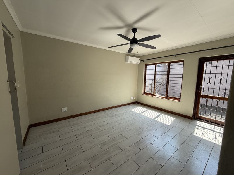 An empty bedroom featuring tiled flooring, a ceiling fan, a wall-mounted air conditioner, and security bars on the windows and door.