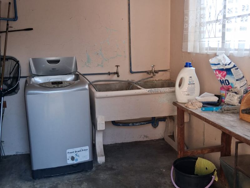 A functional laundry room featuring a double concrete sink, exposed plumbing, and security bars on the window, requiring cosmetic updates to the walls and finishes.