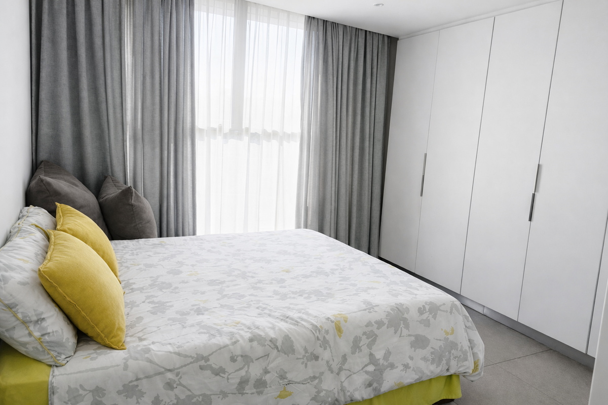 A modern bedroom featuring floor-to-ceiling built-in wardrobes, neutral tiled flooring, and large windows dressed with floor-length curtains.