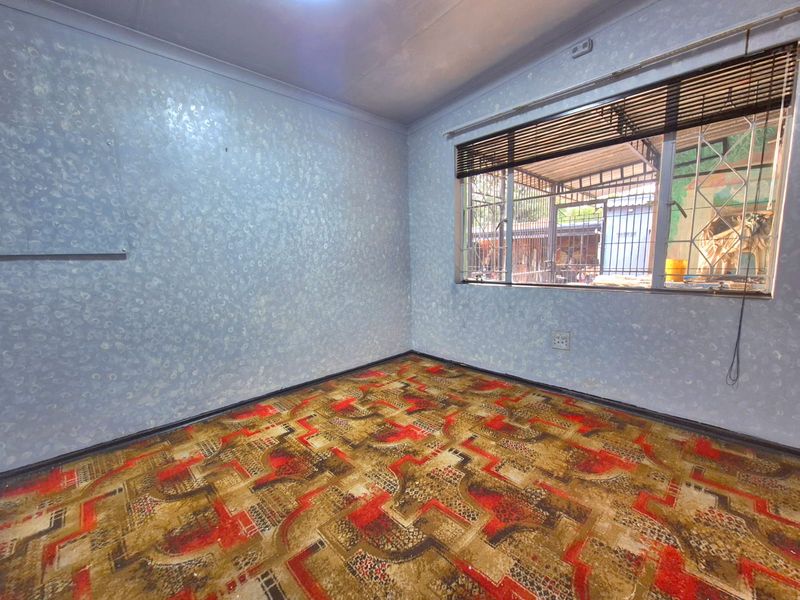 An empty bedroom featuring textured wallpaper, patterned carpeting, and a window fitted with security bars and venetian blinds.