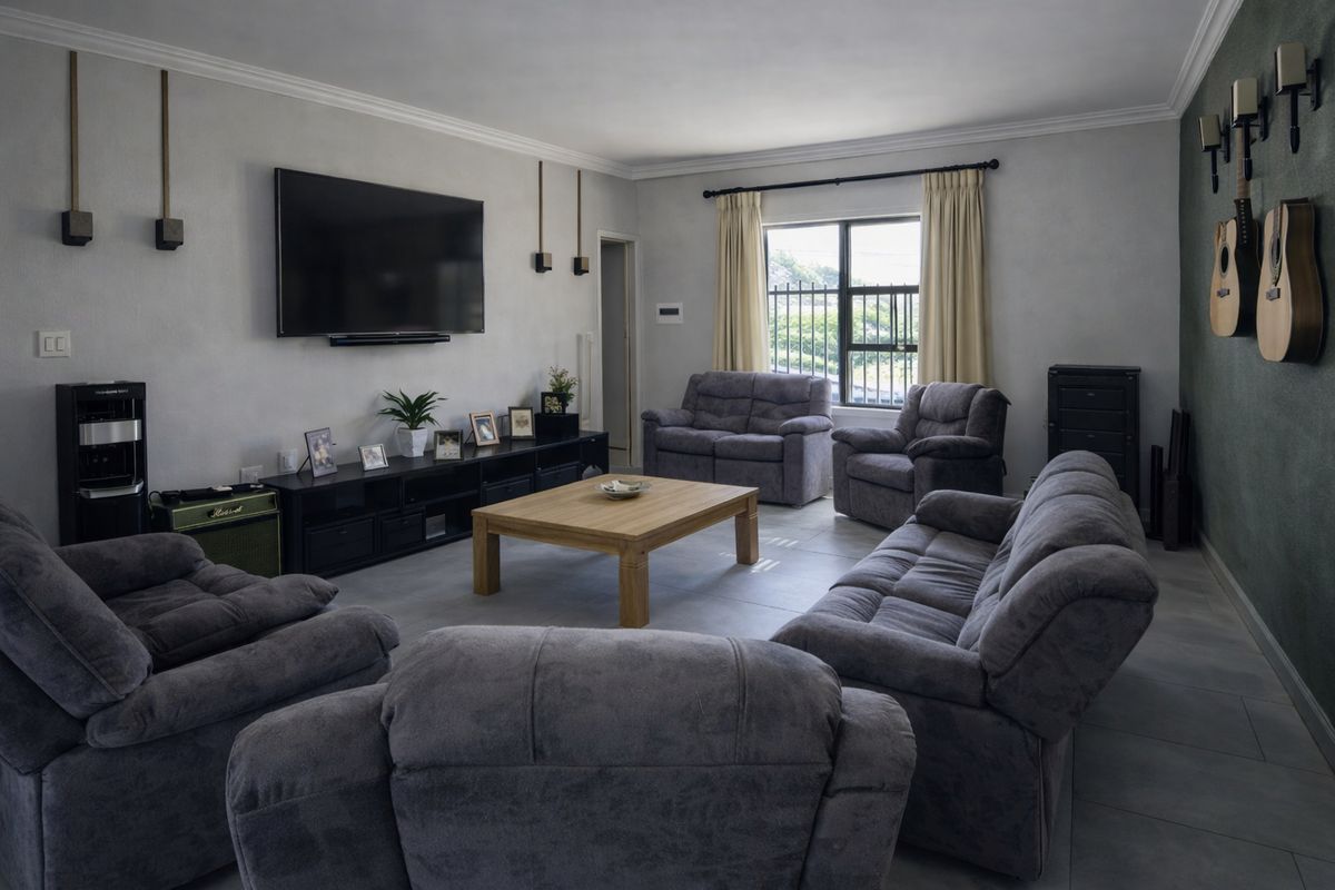 A spacious living room featuring neutral tiled flooring, coved ceilings, and a large window with security bars, providing a comfortable and well-maintained living space.