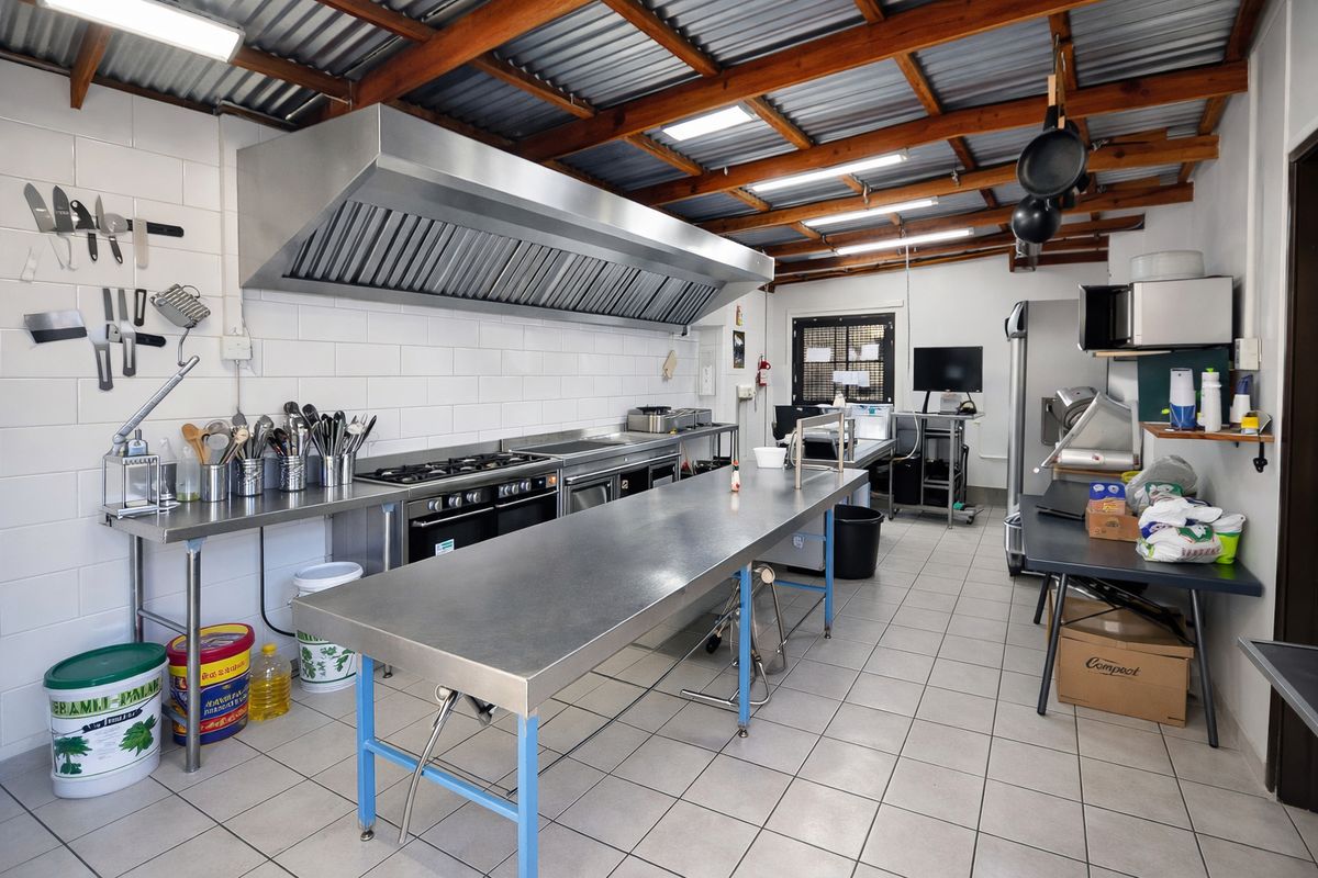 A functional commercial-style kitchen featuring stainless steel prep surfaces, a large extractor hood, gas stove, and exposed timber roof trusses.