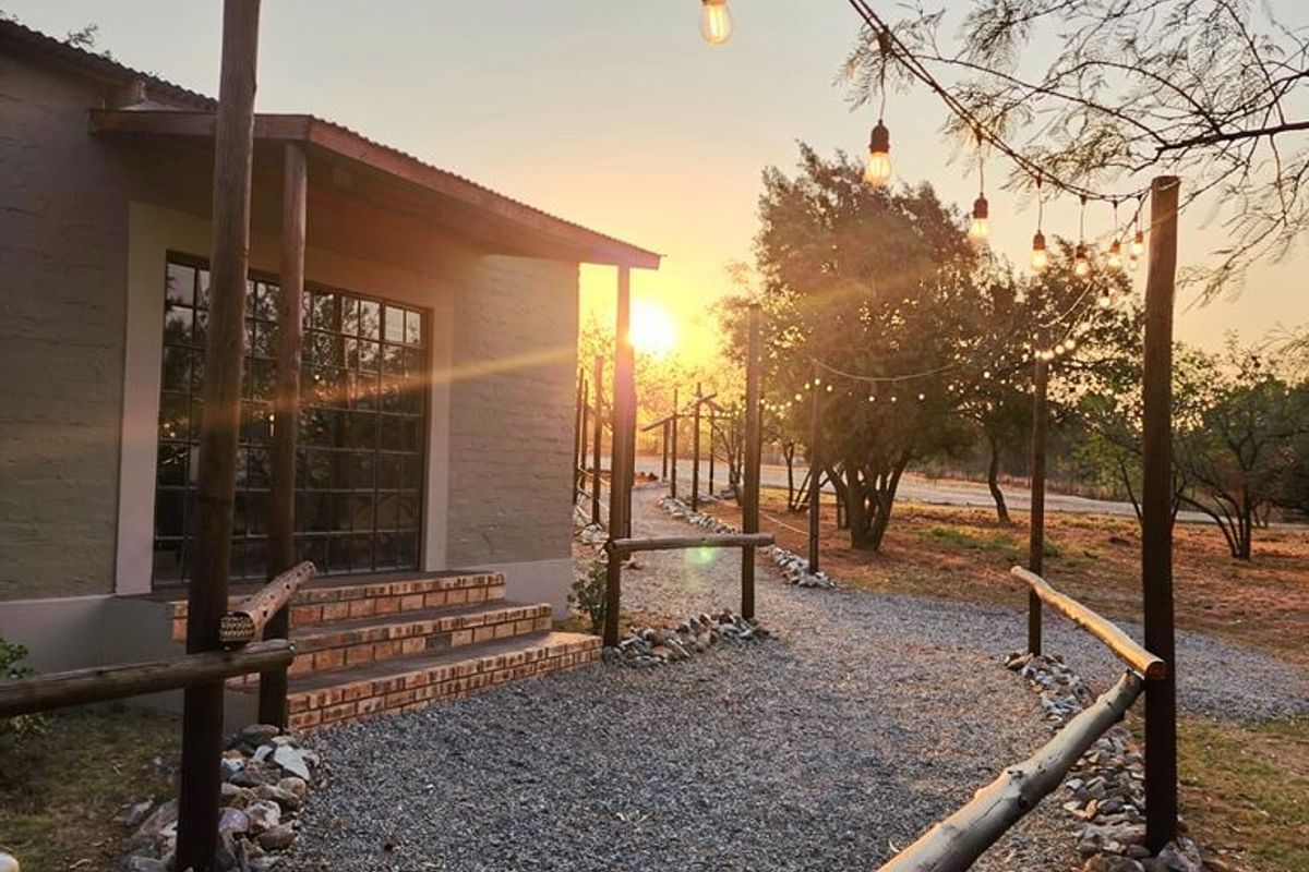 A scenic outdoor area featuring a gravel pathway lined with timber pole fencing, stone borders, and overhead string lighting leading to the building entrance.