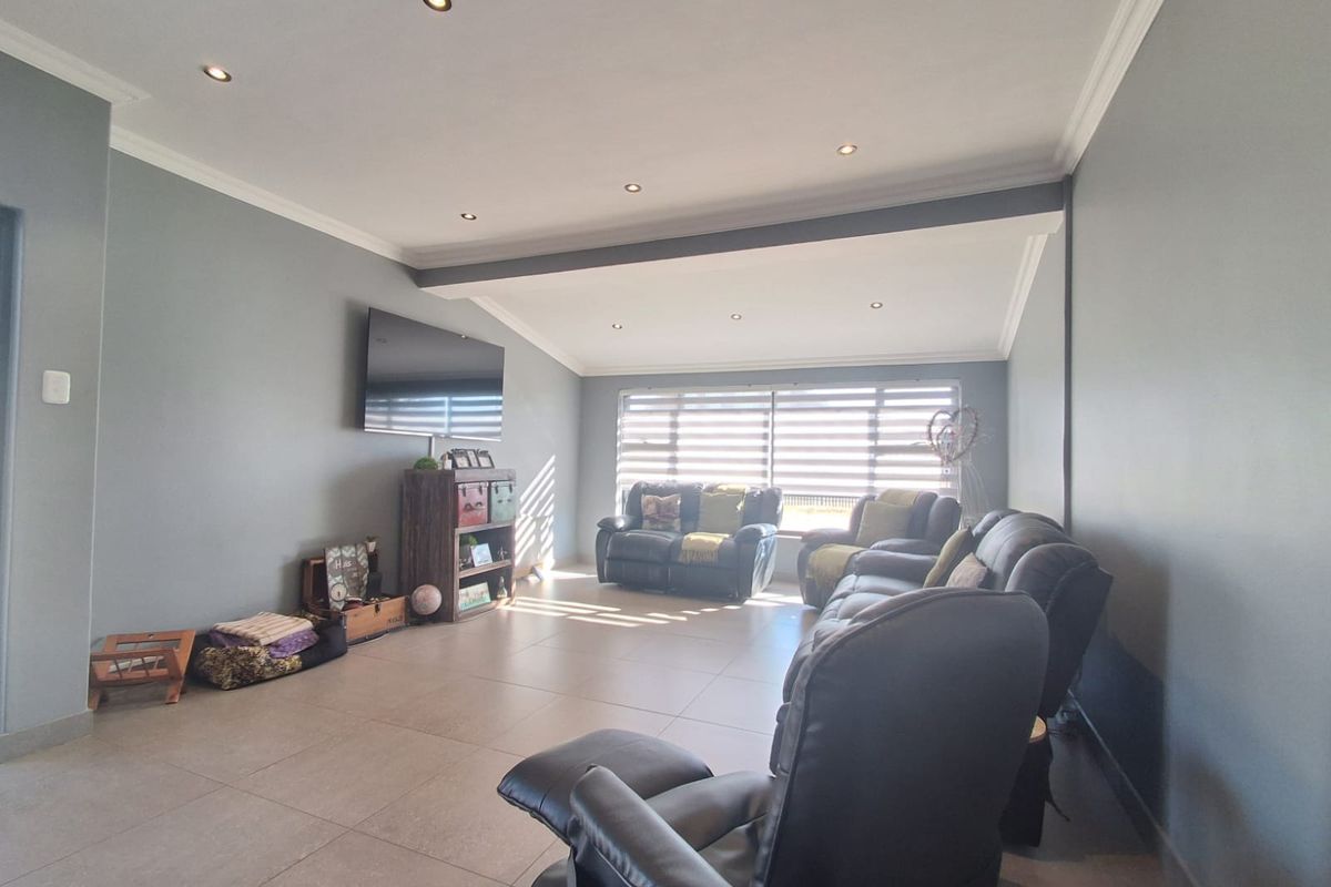 A spacious living room featuring neutral tiled flooring, recessed downlighting, crown molding, and large windows fitted with modern venetian blinds.