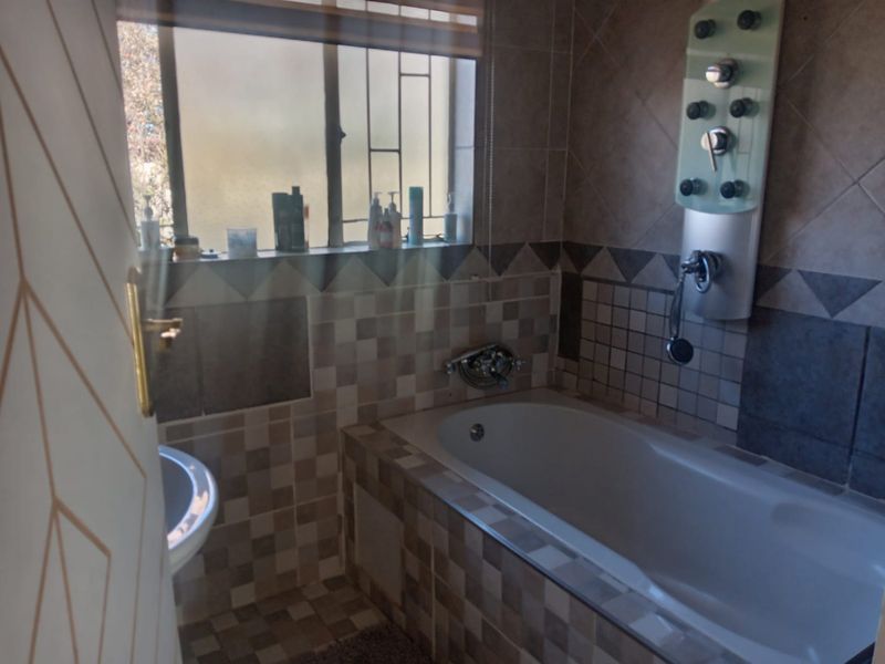 A bathroom featuring a built-in bathtub with a modern shower panel, tiled finishes, and a window with security bars.