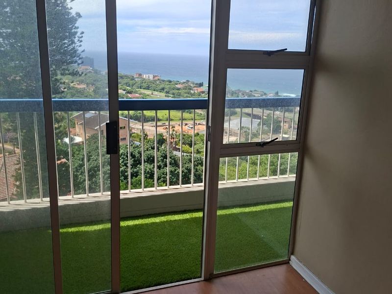 A balcony area featuring artificial grass flooring, a metal balustrade, and sliding glass doors, offering an elevated view of the surrounding landscape and ocean.