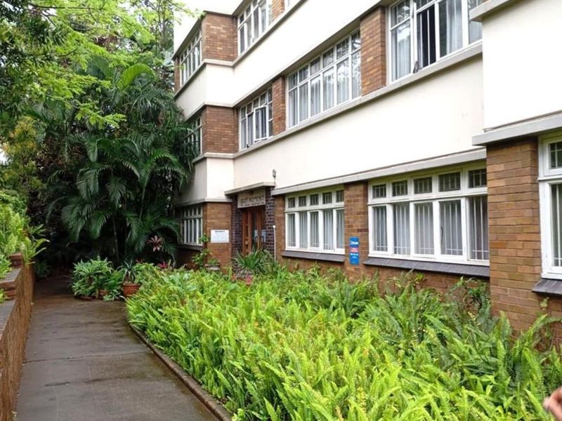 Exterior view of a multi-story apartment building featuring a brick facade, large multi-pane windows, and a paved walkway surrounded by lush garden beds.
