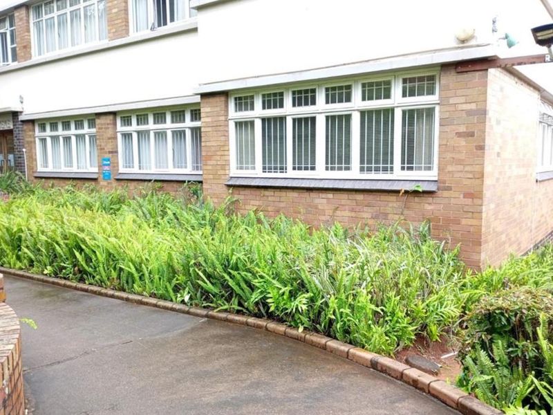 Exterior view of an apartment building featuring a brick facade, multi-pane windows, and a landscaped garden bed along a concrete pathway.
