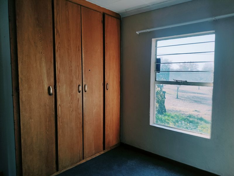 A bedroom featuring built-in wooden cupboards and a window with security bars, offering a view of the exterior grounds.