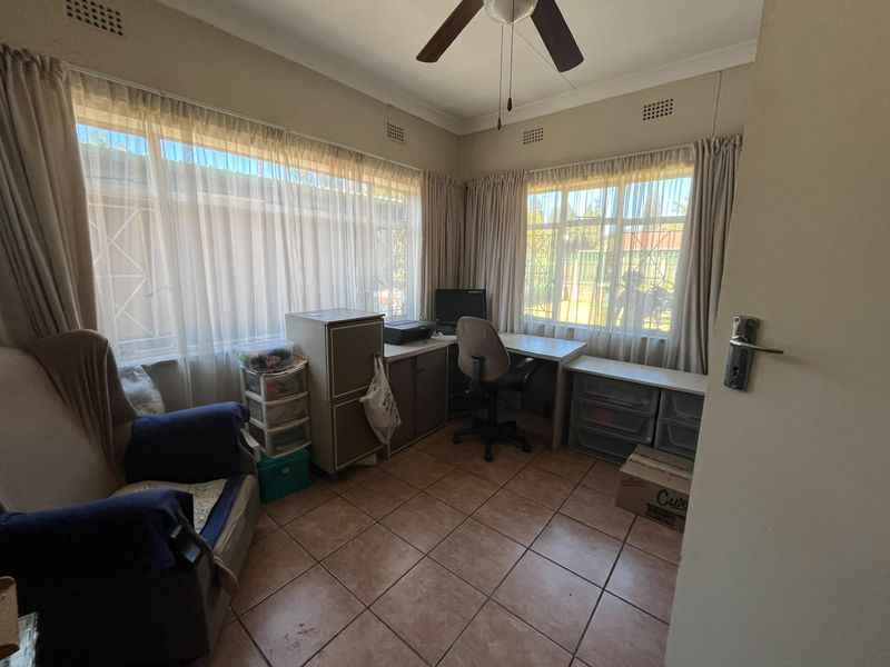 A functional home office space featuring tiled flooring, a ceiling fan, and large windows with security bars, offering natural light and a practical layout.