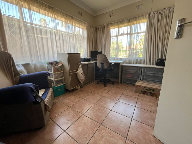 A functional home office space featuring tiled flooring, large windows with security bars, and neutral wall finishes.