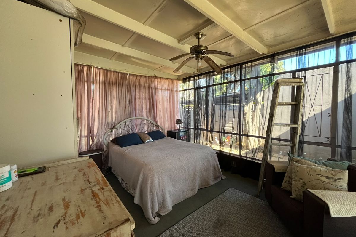 A bedroom featuring an exposed beam ceiling, ceiling fan, and large windows with security bars, offering natural light and garden views.