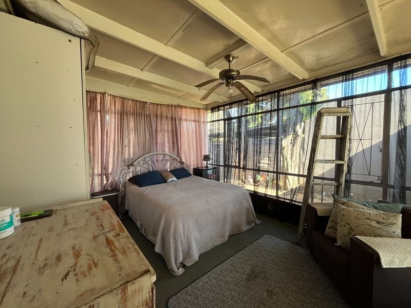 A bedroom featuring an exposed beam ceiling, ceiling fan, and large windows with security bars, offering natural light and garden views.