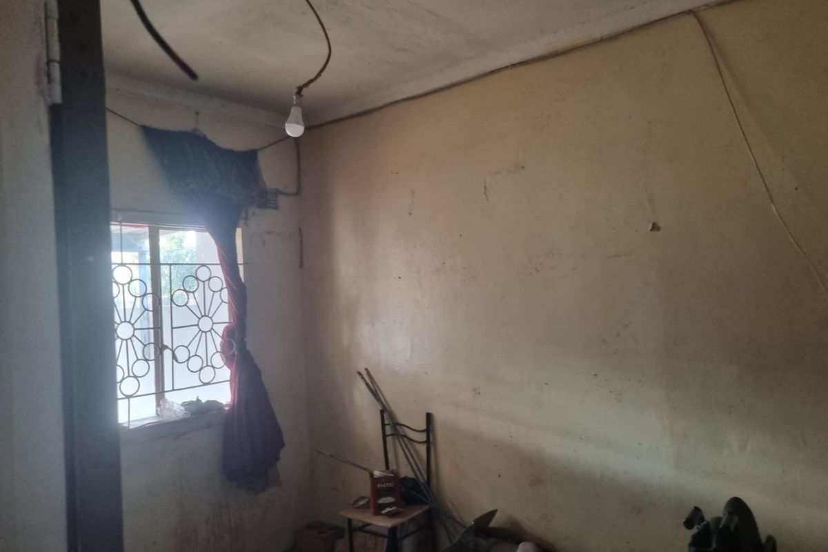 A bedroom in need of significant renovation, featuring exposed electrical wiring, damaged wall finishes, and a security gate on the window.