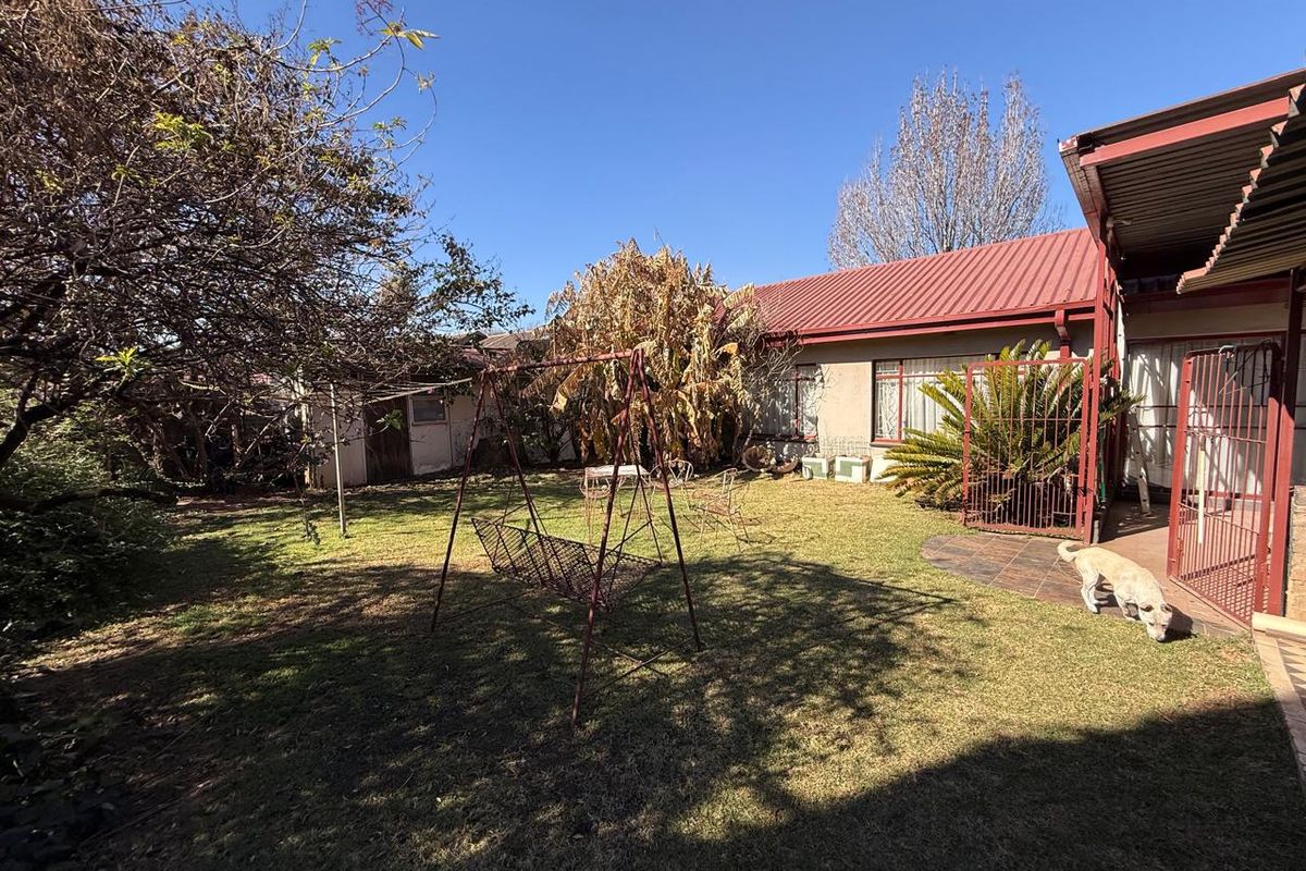 Rear garden area featuring a lawn, mature trees, and a covered patio section with security gate access to the main house structure.