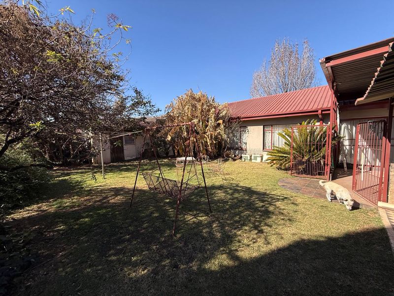 Rear garden area featuring a lawn, mature trees, and a covered patio section with security gate access to the main house structure.