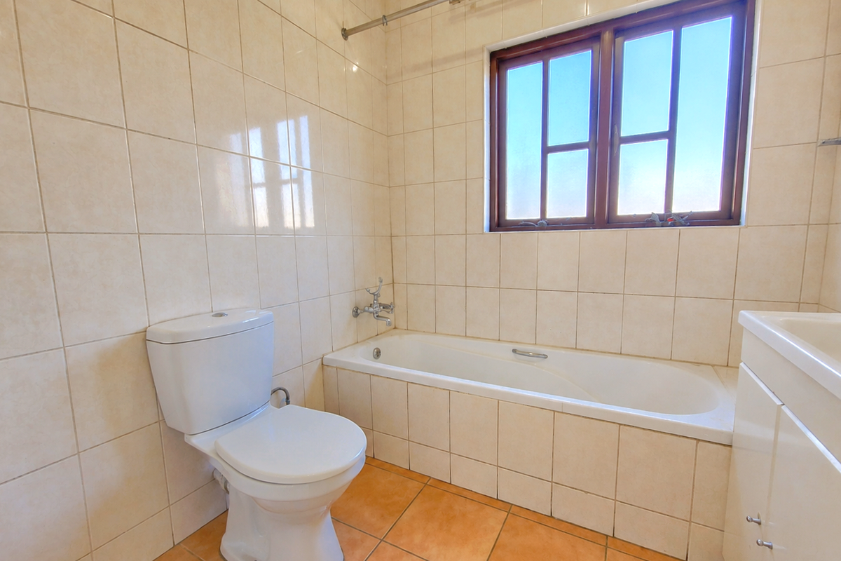 A functional bathroom featuring a built-in bathtub, toilet, and vanity cabinet with neutral wall tiling and terracotta-style floor tiles.