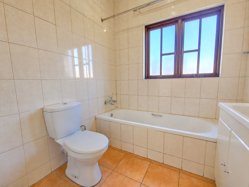 A functional bathroom featuring a built-in bathtub, toilet, and vanity cabinet with neutral wall tiling and terracotta-style floor tiles.