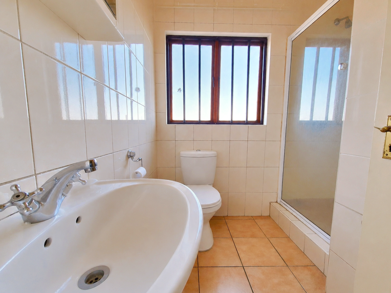 A functional bathroom featuring a pedestal basin, toilet, and a glass-enclosed shower with neutral wall and floor tiling and security bars on the window.