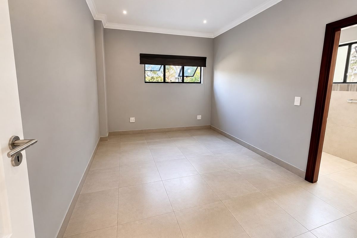 A clean, neutral-toned bedroom featuring tiled flooring, recessed lighting, and a window with a roller blind, offering a blank canvas for personalization.