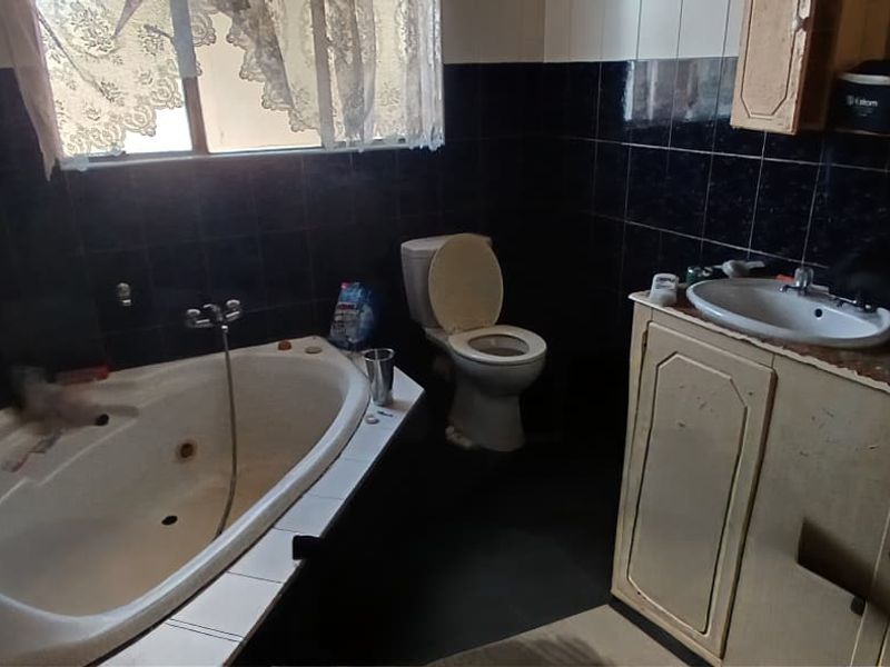 A dated bathroom featuring a corner bathtub, dual vanity unit, and toilet, finished with dark wall tiles.