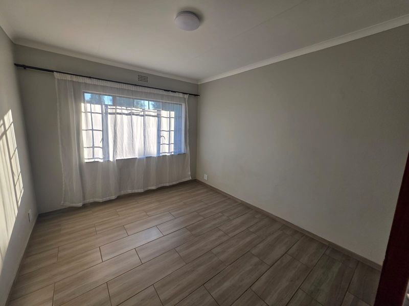 A clean, empty bedroom featuring neutral wall tones, modern wood-look tiled flooring, and a window fitted with security bars.