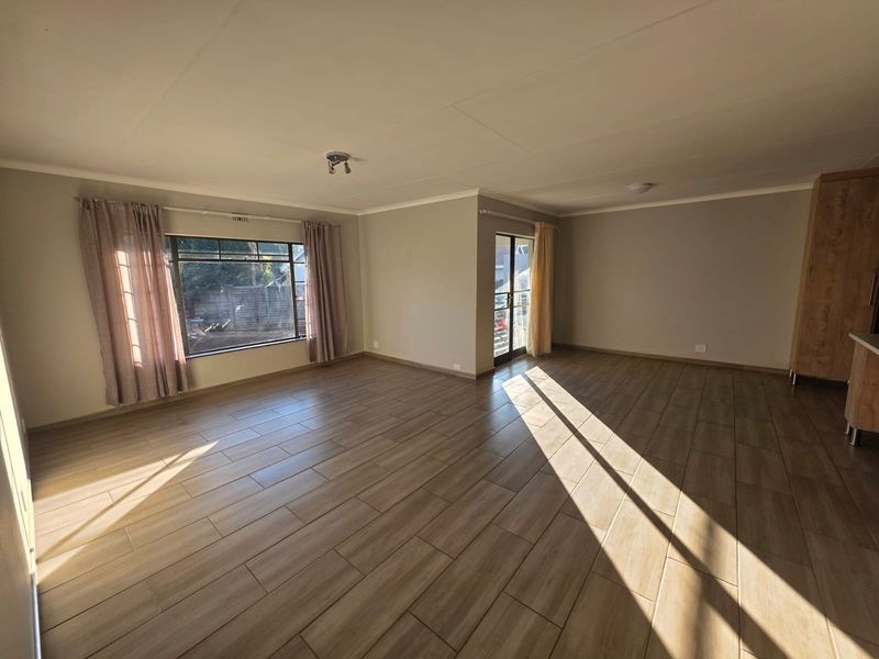 Spacious open-plan living area featuring modern wood-look tiled flooring, neutral walls, and abundant natural light through large windows and a glass door.