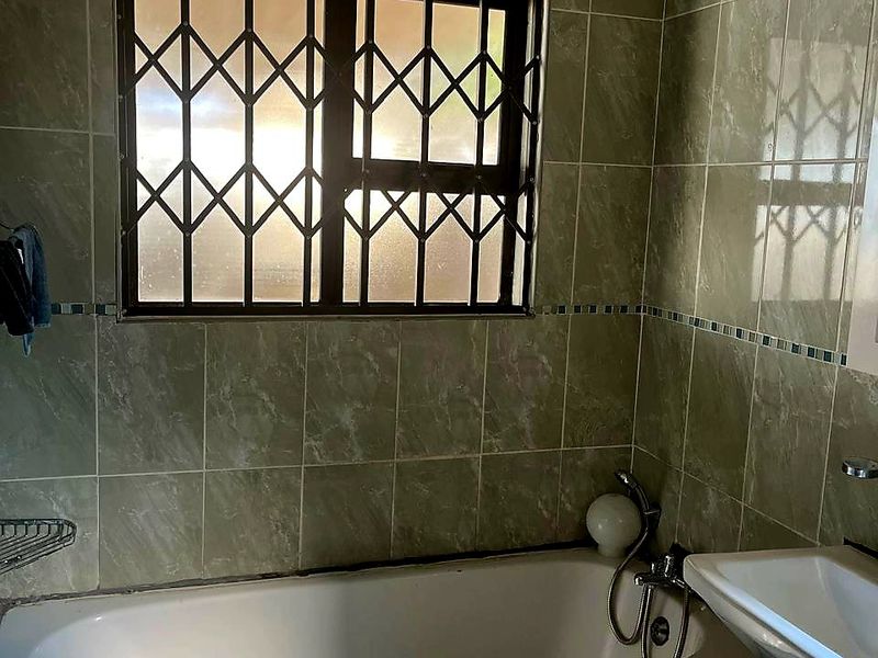 A dated bathroom featuring a bathtub, pedestal basin, and full-height wall tiling with a security gate on the window.