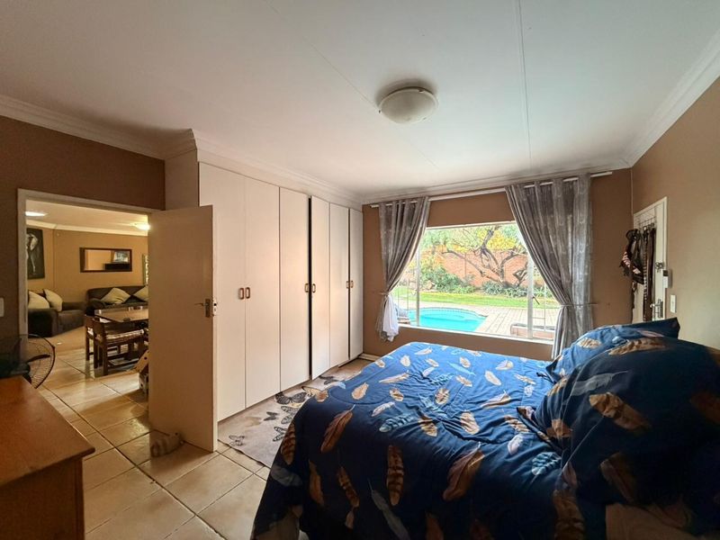 Spacious bedroom featuring built-in cupboards, tiled flooring, and a large window overlooking the garden and swimming pool area.