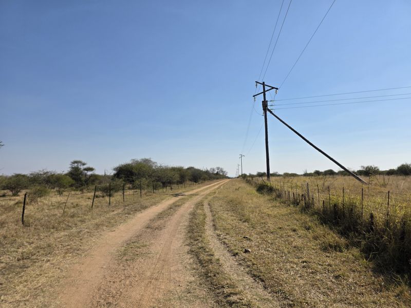 A rural dirt access road flanked by wire fencing and utility infrastructure, characteristic of open farm land.