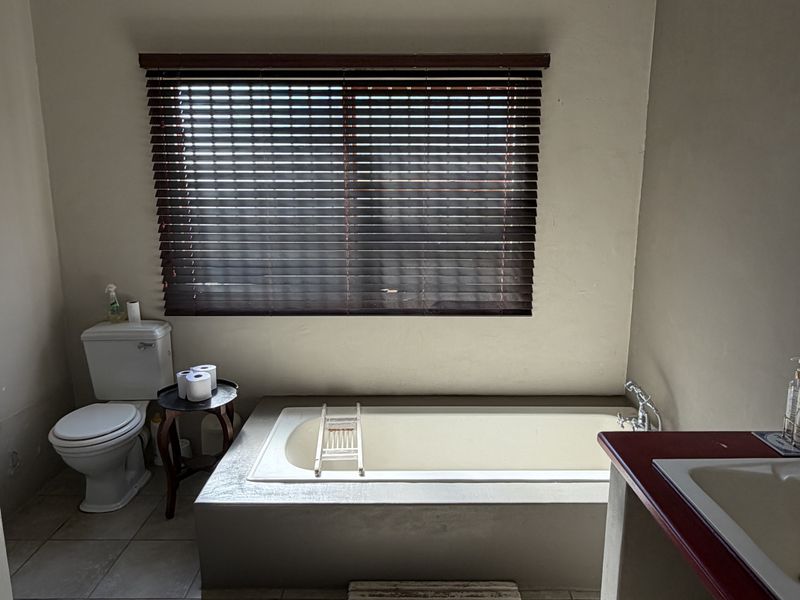 A functional bathroom featuring a built-in bathtub, toilet, and vanity basin with tiled flooring and timber blinds.