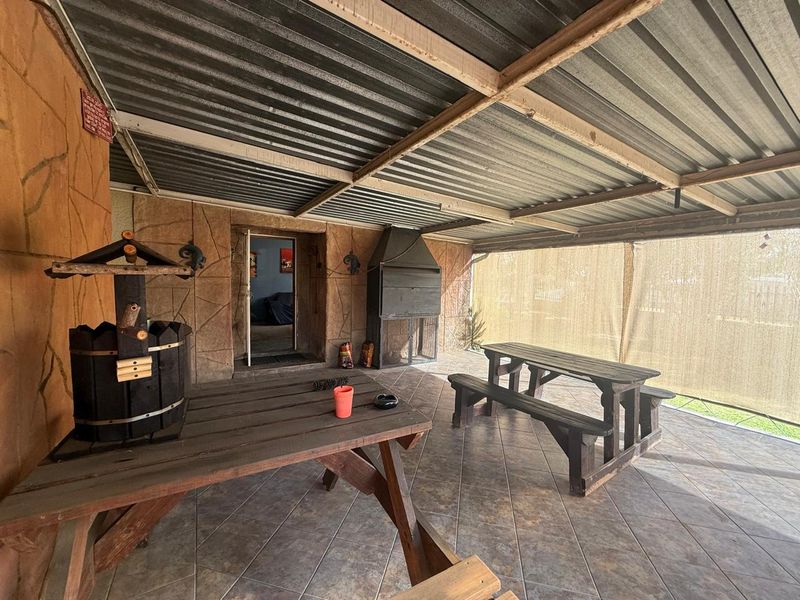 Covered outdoor patio area featuring tiled flooring, a built-in braai, and a corrugated metal roof structure, suitable for outdoor entertaining.