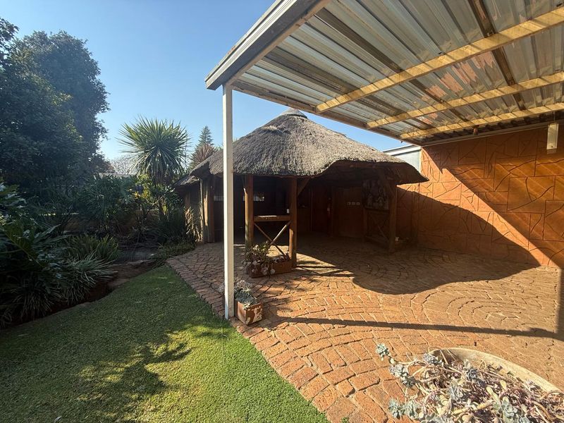 Outdoor entertainment area featuring a traditional thatched lapa, covered patio with brick paving, and a textured feature wall in a landscaped garden setting.