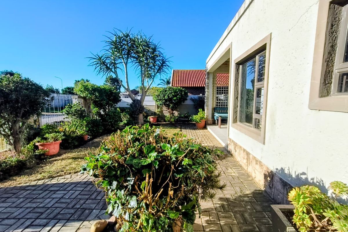 A rear exterior view of the property featuring a paved patio area, established garden landscaping, and plastered exterior walls with aluminium window frames.