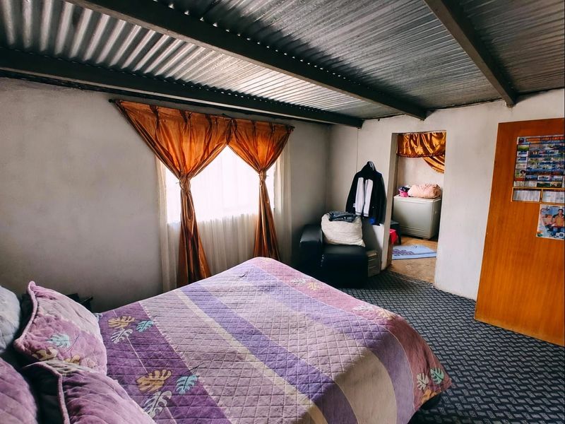 A bedroom featuring a corrugated metal ceiling, patterned carpet flooring, and a simple timber door, offering a basic living space.