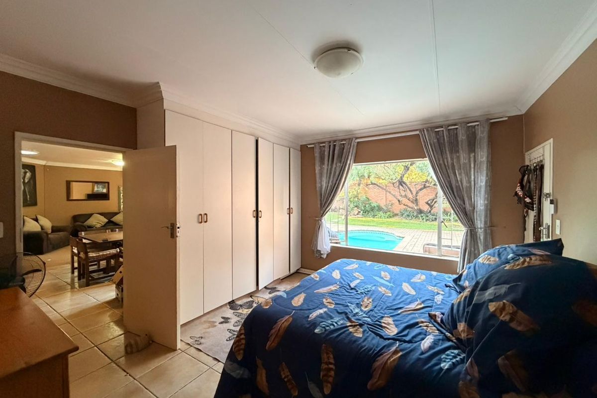 Spacious bedroom featuring built-in cupboards, tiled flooring, and a large window overlooking the garden and swimming pool area.