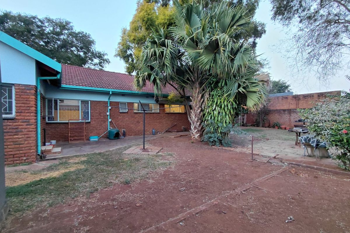 Rear view of the house featuring a brick facade, tiled roof, and a spacious backyard with a large palm tree and paved patio area.
