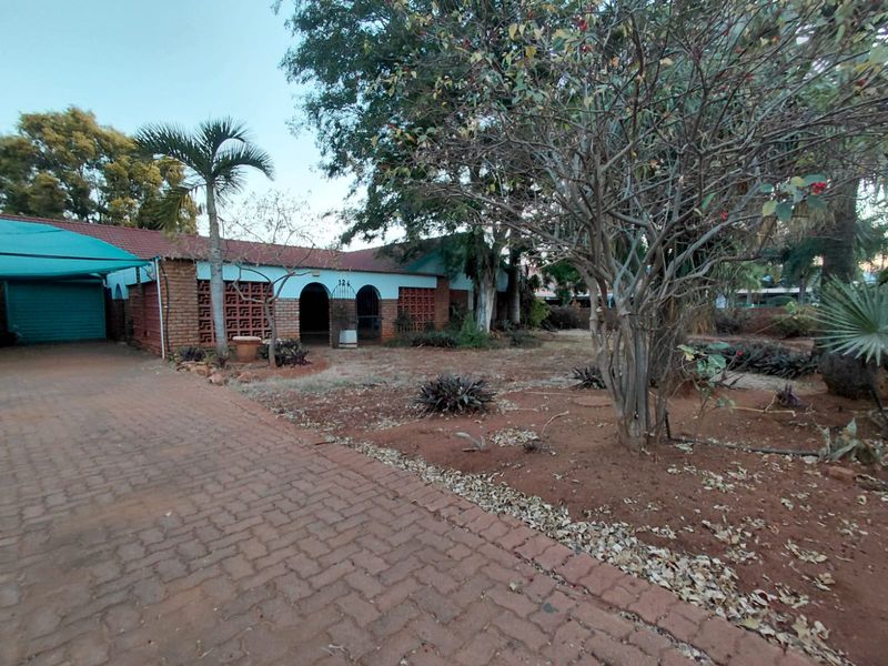 Front exterior view of a single-story house featuring a brick driveway, carport, and established garden with mature trees and breeze block architectural details.