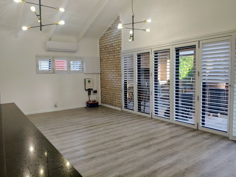 Spacious open-plan living area featuring vaulted ceilings, exposed brickwork, modern laminate flooring, and large bi-fold doors with plantation shutters opening to the outdoors.