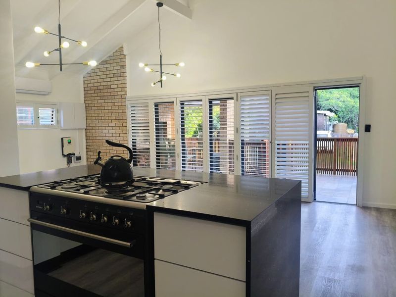 Modern kitchen featuring a central island with gas stove, vaulted ceilings, exposed brick accents, and plantation shutters opening to an outdoor deck.