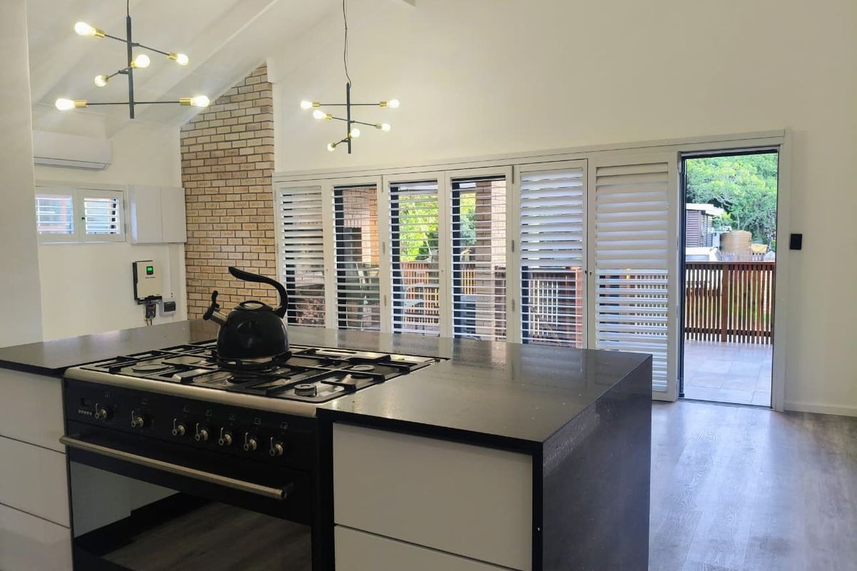 Modern kitchen featuring a central island with gas stove, vaulted ceilings, exposed brick accents, and plantation shutters opening to an outdoor deck.