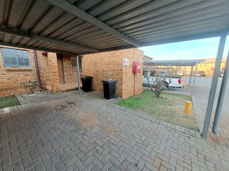 A covered carport area with facebrick exterior walls and paved driveway, featuring a fire hose reel and small garden patch.