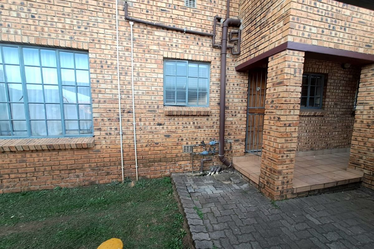 Rear exterior view of a face brick townhouse featuring a paved patio area, security gate, and visible external plumbing infrastructure.