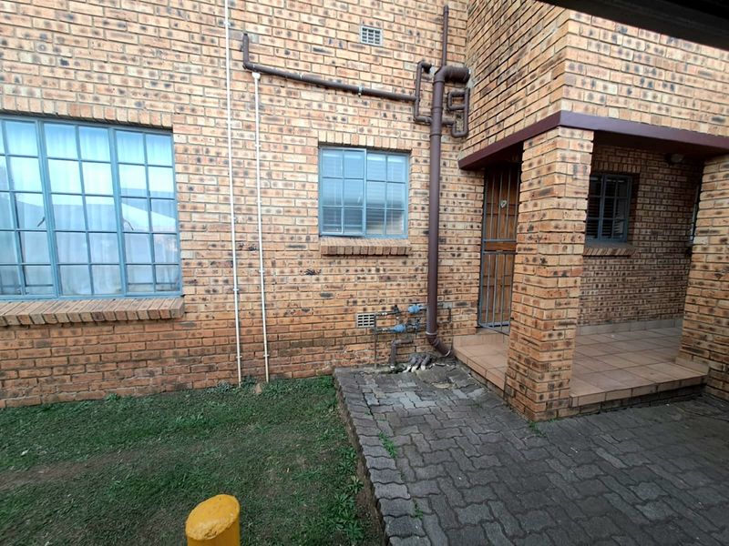 Rear exterior view of a face brick townhouse featuring a paved patio area, security gate, and visible external plumbing infrastructure.