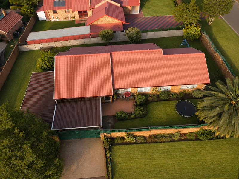 Aerial view of a residential property featuring a large corrugated metal roof, paved driveway, secure perimeter wall, and a well-maintained garden.