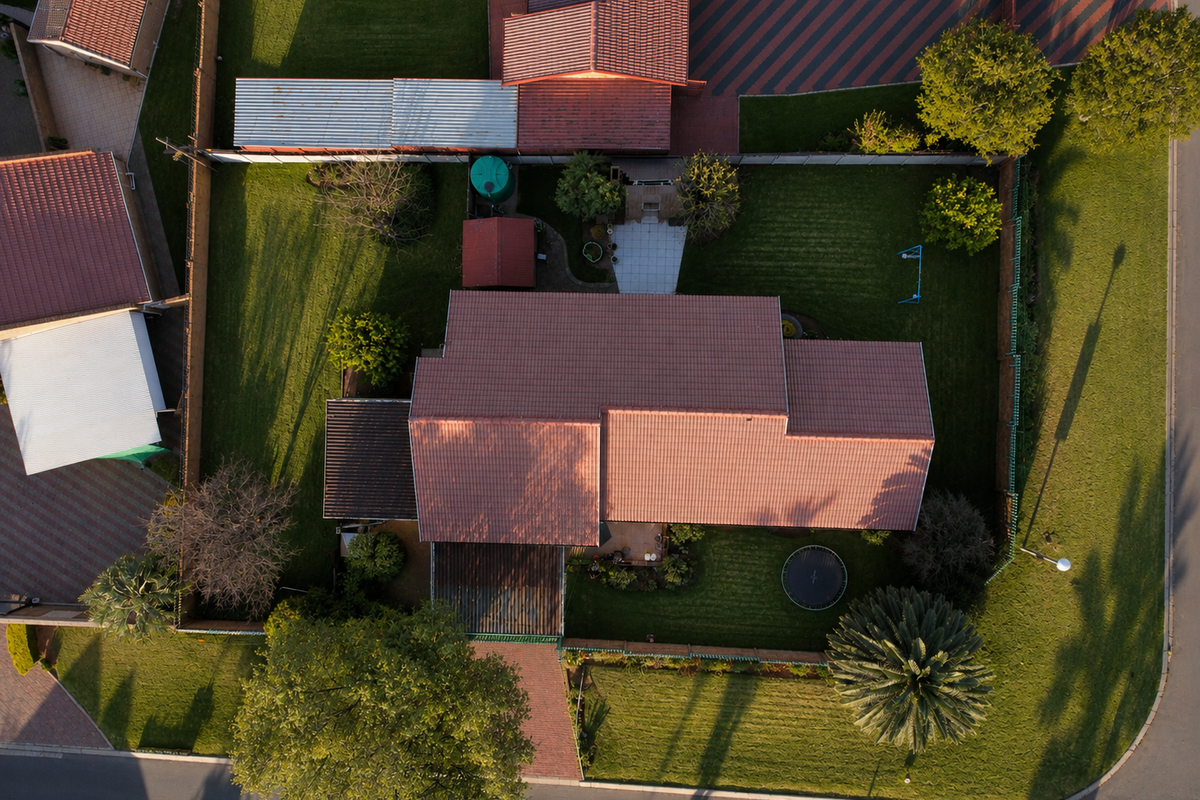 Aerial view of the property showcasing the roof structure, paved driveway, expansive lawn, and established garden areas within the fenced perimeter.